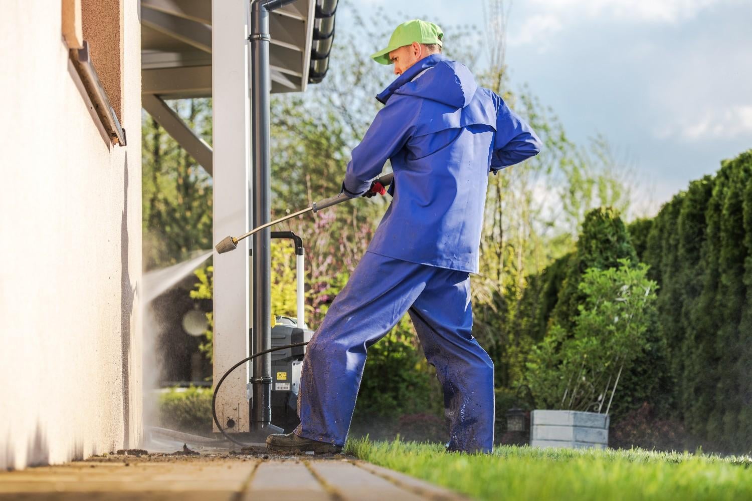 A person in blue waterproof gear and a green cap power washing a house exterior wall and the ground.