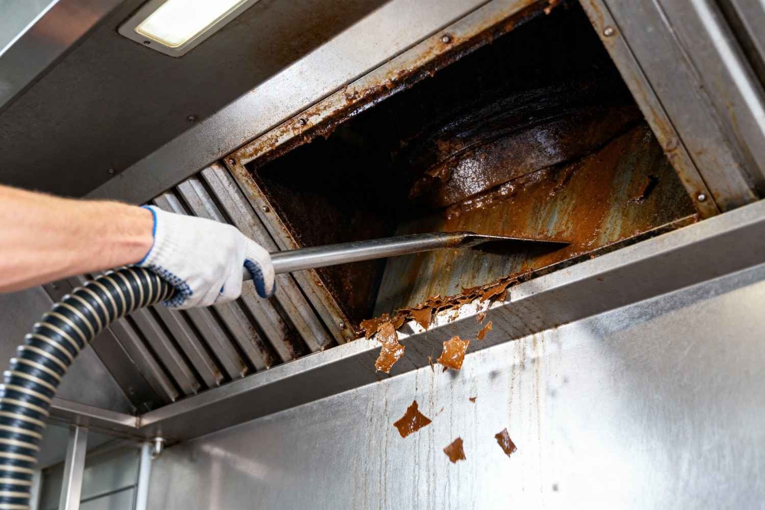 A gloved hand uses a scraper and vacuum hose to remove thick, congealed grease from a grimy industrial kitchen exhaust hood.