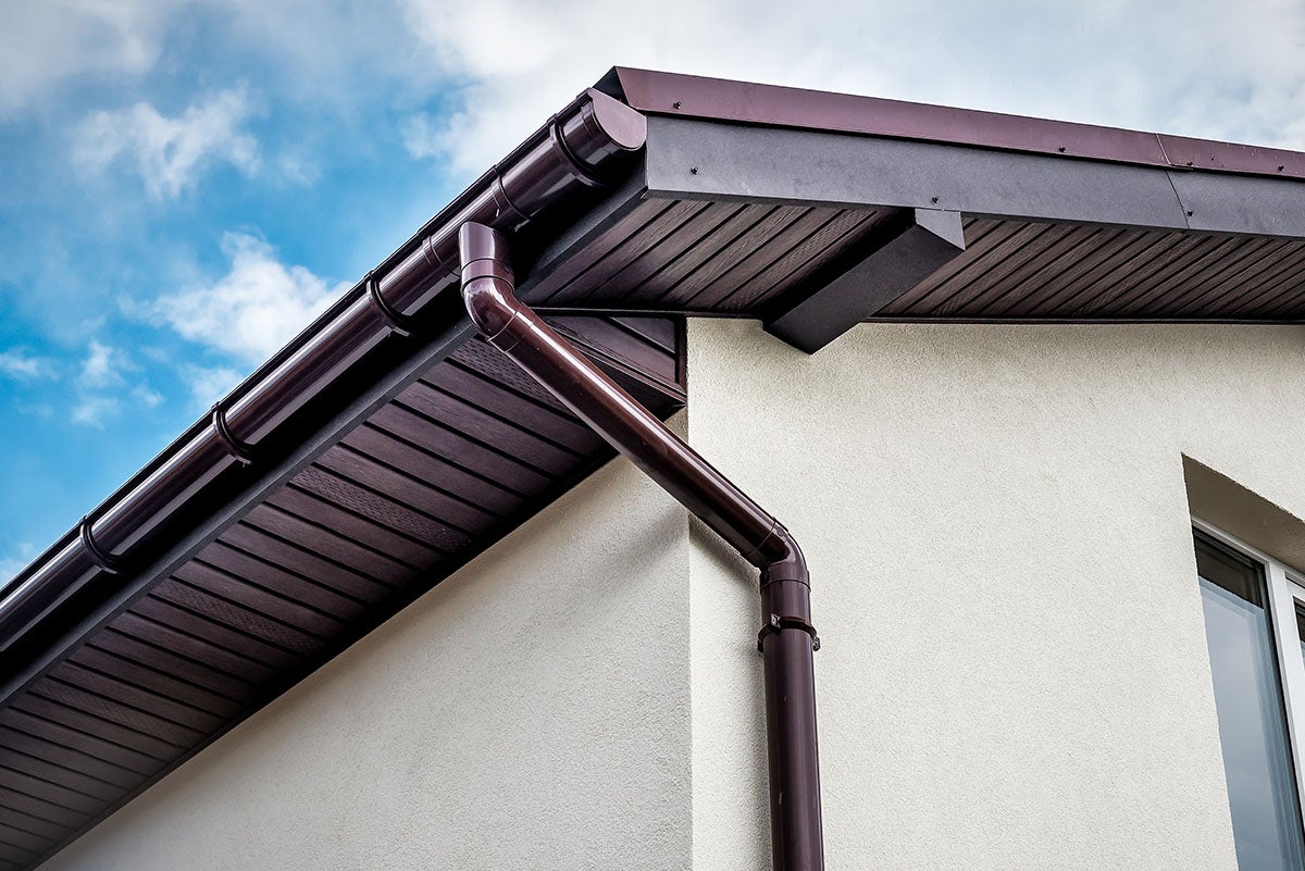 Brown gutters and downspout on a light-colored building with a dark brown facade against a blue and cloudy sky.